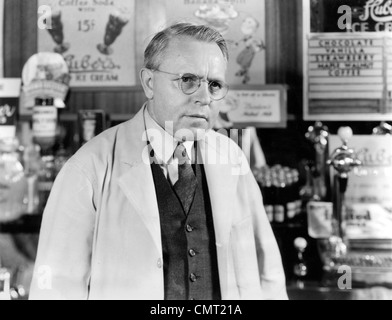 1930S 1940S DROGERIE SODABRUNNEN MANN APOTHEKER TRAGEN WEIßEN MANTEL UND BRILLE HÖREN Stockfoto