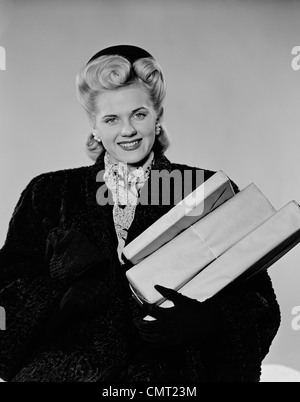 1940ER JAHRE BLONDE FRAU LÄCHELND TRAGEN WINTER MANTEL TRAGEN SHOPPING PAKET BLICK IN DIE KAMERA Stockfoto