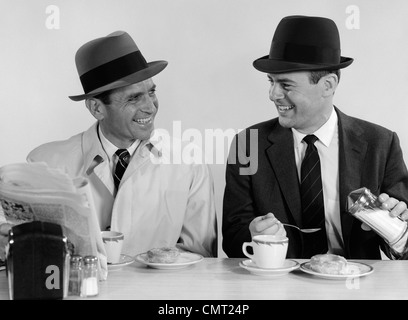 1960ER JAHRE ZWEI GESCHÄFTSLEUTE TRAGEN HÜTE, TRINKEN KAFFEE LÄCHELN SPRECHEN SITZEN ZUSAMMEN AN EINEM DINER MITTAGESSEN SCHALTER Stockfoto