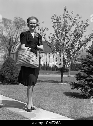 1930S 1940S STILVOLL GEKLEIDETE FRAU AUF SUBURBAN STRAßE STROH TRAGETASCHE Stockfoto