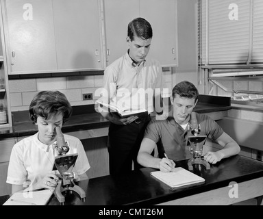 1960ER JAHREN DREI STUDENTEN, DIE MIT MIKROSKOPEN ZWEI JUNGEN EIN MÄDCHEN INNEN HIGH SCHOOL Stockfoto