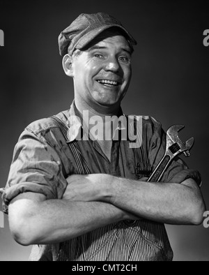 1950S 1960S LÄCHELND PORTRAIT MANN IM GESTREIFTEN CAP & OVERALLS ÜBERQUEREN ARME SCHRAUBENSCHLÜSSEL BLICK IN DIE KAMERA HALTEN Stockfoto