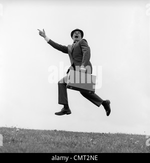 1960ER JAHREN GESCHÄFTSMANN MIT EINER AKTENTASCHE LAUFEN SCHREIEN UND SPRINGEN IN DIE LUFT IM FREIEN ZEIGT FINGER NACH VORNE Stockfoto