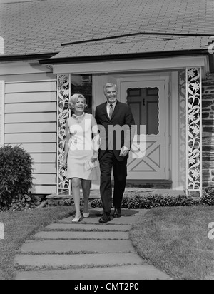 1960ER JAHRE ÄLTERES PAAR ZU FUß AUF DEM BÜRGERSTEIG VOR DEM HAUS Stockfoto