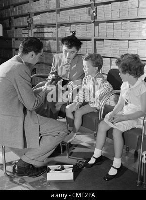 1950ER JAHRE FAMILIE MUTTER ZWEI KINDER IM SCHUHGESCHÄFT VERSUCHEN NEUE SCHUHE GEHOLFEN DURCH VERKÄUFER Stockfoto