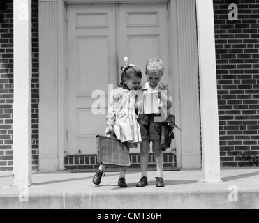 1940ER JAHRE KLEINE JUNGEN UND MÄDCHEN MIT SCHULRANZEN BLICK AUF NOTEPAD IM FREIEN Stockfoto