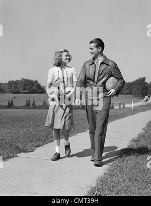 1930S 1940S TEEN PAAR HINUNTER BÜRGERSTEIG MÄDCHEN MIT SCHULJUNGE BÜCHER HALTEN FUßBALL Stockfoto