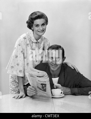 1960ER JAHRE MANN FRAU TRAGEN PYJAMA AM FRÜHSTÜCK TISCH FRAU HINTER MANN HALTEN ZEITUNG BEIDE LÄCHELND UND BLICK IN DIE KAMERA Stockfoto