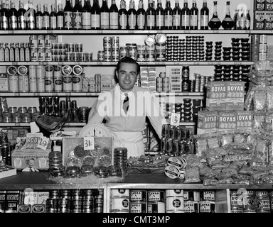 1940ER JAHREN LÄCHELND MANN LEBENSMITTELHÄNDLER STEHEN HINTER THEKE GEFÜLLT MIT VERSCHIEDENEN LEBENSMITTELN IN DOSEN UND PAKETE INDOOR LEBENSMITTELGESCHÄFT Stockfoto