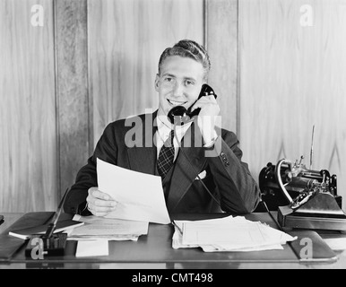 1930S 1940S LÄCHELND GESCHÄFTSMANN GESPRÄCH AM TELEFON SITZEN AM SCHREIBTISCH MIT DIKTIERGERÄT Stockfoto