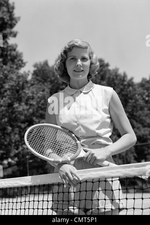 1950ER JAHRE PORTRAIT FRAU MIT TENNISSCHLÄGER HINTER NETTO IM FREIEN BLICK IN DIE KAMERA Stockfoto