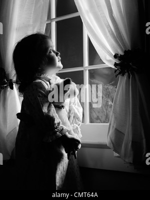 1950ER JAHRE MÄDCHEN IN FLANELL-NACHTHEMD MIT AUSGESTOPFTEN TIER BLICK AUS FENSTER IN DER NACHT MIT VORHÄNGEN HELD ZURÜCK VON HOLLY Stockfoto