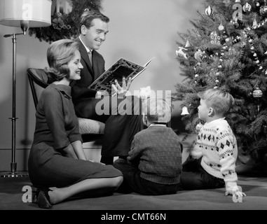 1960ER JAHRE BAUM FAMILIE VATER MUTTER ZWEI SÖHNE LÄCHELND BIS WEIHNACHTEN SITZEN IM WOHNZIMMER, EIN BUCH ZU LESEN Stockfoto