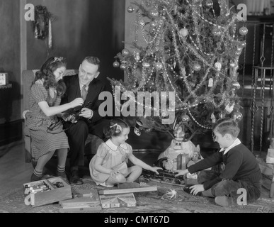 1930ER JAHRE FAMILIE WEIHNACHTEN FOTO VATER, ZWEI TÖCHTER UND SOHN MIT GESCHENKEN UND BAUM Stockfoto