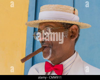 Ein dunkle kubanischen älterer Herr im weißen Anzug, Hut Stroh Bootsfahrer, rote Fliege und eine Zigarre zeichnet sich durch eine bunte Wand in Havanna. Stockfoto