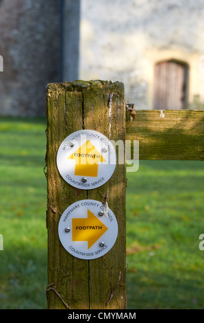 Hampshire County Council Wanderweg Markierung oder Richtung Zeichen auf einem alten Holzzaun mit einer Dorfkirche im Hintergrund. Stockfoto