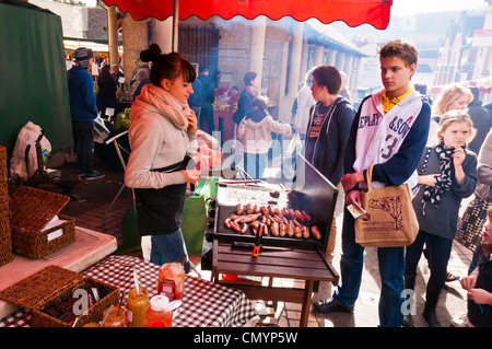 Junge Frau Standinhaber Kochen Würstchen am Samstag Bauernmarkt in Stroud, Gloucestershire, UK Stockfoto