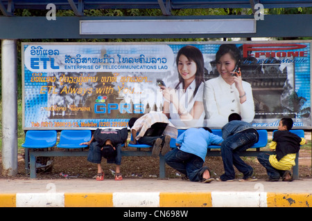 Eine Gruppe von jungen spielen an einer Bushaltestelle unterhalb einer Telekommunikation Firmenschild auf einer Stadtstraße in Vientiane, Laos. Stockfoto