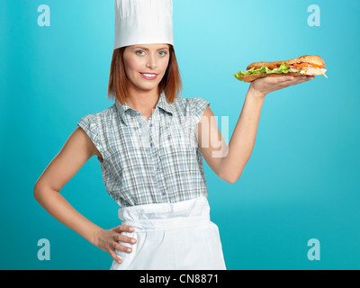 Porträt der schönen jungen Frau Koch, hält einen großen Sandwich in einer ihrer Hände auf blauem Hintergrund Stockfoto