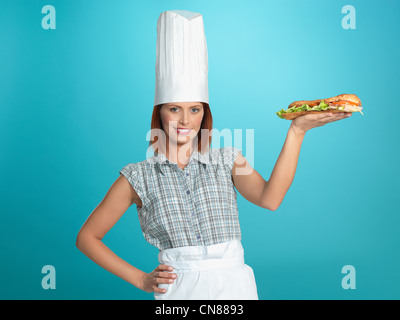 Porträt der schönen jungen Frau Koch, hält einen großen Sandwich in einer ihrer Hände auf blauem Hintergrund Stockfoto