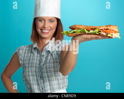 Porträt der schönen jungen Frau Koch, hält einen großen Sandwich auf einer ihrer Hände auf blauem Hintergrund Stockfoto