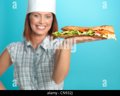 Porträt der schönen jungen Frau Koch, hält einen großen Sandwich in einer ihrer Hände auf blauem Hintergrund Stockfoto