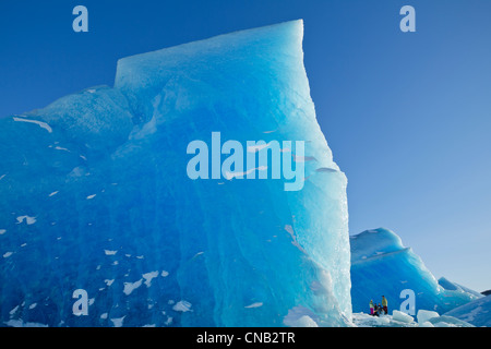 Eine Wand aus blauen Eis überragt Wanderer, wie sie einen riesigen Eisberg eingefroren in die Oberfläche des Mendenhall Lake, Alaska erkunden Stockfoto