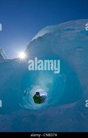 Zusammensetzung: Person, die durch einen kleinen Tunnel erodiert in einem Eisberg eingefroren in Mendenhall Lake, Juneau, Alaska, Winter Stockfoto