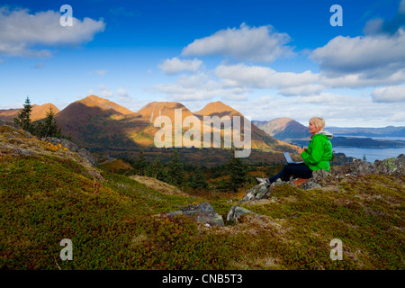 Reife Frau Typen auf Laptop-Computer sitzend auf Säule Berg mit Monashka Bucht im Hintergrund, Kodiak Island, Alaska Stockfoto