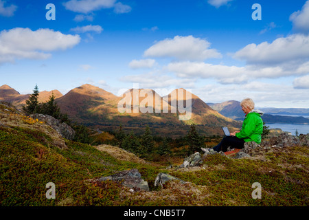 Reife Frau Typen auf Laptop-Computer sitzend auf Säule Berg mit Monashka Bucht im Hintergrund, Kodiak Island, Alaska Stockfoto