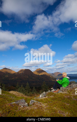 Reife Frau entspannt und genießt ein Heißgetränk auf Säule Berg mit Monashka Bucht im Hintergrund, Alaska, Kodiak Insel, Südwesten Stockfoto