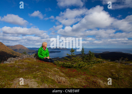 Reife Frau entspannt und schreibt im Journal auf Säule Berg mit Monashka Bucht im Hintergrund, Kodiak Insel, Alaska, Herbst Stockfoto