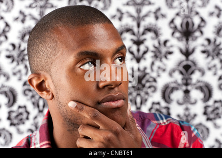 Ein junger afrikanischer Amerikaner Mann in seinen Zwanzigern und seine Hand auf seinem Kinn denken tief über etwas vor einem Damast Stil Stockfoto
