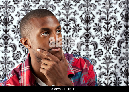 Ein junger afrikanischer Amerikaner Mann in seinen Zwanzigern und seine Hand auf seinem Kinn denken tief über etwas vor einem Damast Stil Stockfoto