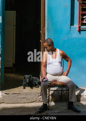 Ein älterer kubanischer erwachsener Mann sitzt auf einem Stuhl vor der offenen Tür seines Hauses und befasst sich mit seinem Hund in Havanna, Kuba. Stockfoto