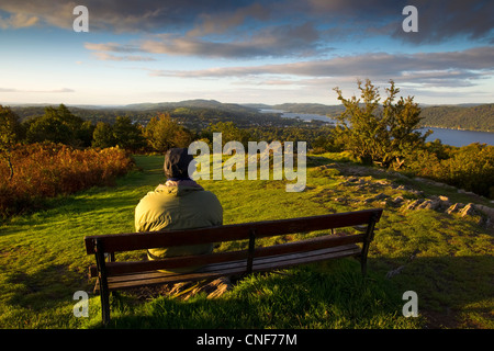 Eine Person mit einer grünen Jacke, die auf einer Bank sitzen auf einem Hügel mit Blick über die Hügel und ein See mit Wolken im Himmel. Stockfoto