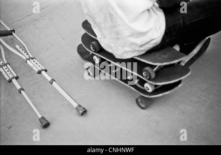 Ein Mann sitzt auf einem Stapel von Skateboards neben Krücken Stockfoto