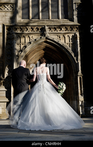 Braut am Tag der Hochzeit von hinten zu Fuß in die Kirche hält Vater/Arm mit großen Kleid mit Rücken und Bouquet geschnürt Stockfoto