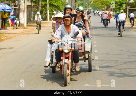 Horizontale Porträt einer vietnamesischen Frau teilen eine Fahrt entlang einer viel befahrenen Straße, ein Motorrad und Anhänger tragen westliche Touristen. Stockfoto