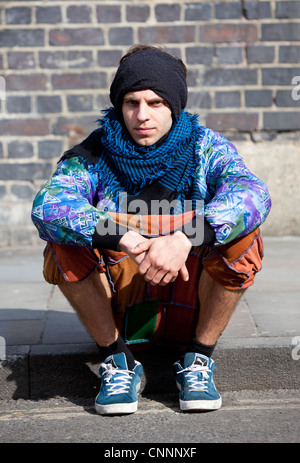 Porträt eines trendigen jungen Mannes auf einer Bordsteinkante, Brick Lane, London, England, Großbritannien. Stockfoto