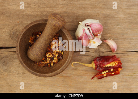 Chili-Flocken in einem Stößel und Mörser mit Knoblauch und geschnittenen Chilischote auf einem alten verwitterten Holzbrett Stockfoto