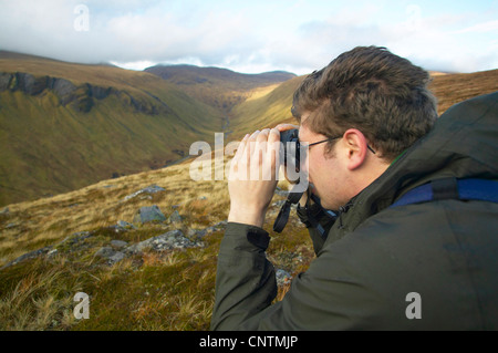 Stalker sitzt auf einem Felsen, Blick auf Tal des Flusses durch ein Fernglas, Alladale Wilderness Reserve, Sutherland, Schottland, Vereinigtes Königreich Stockfoto