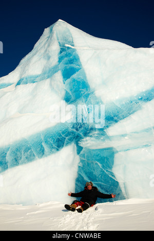 lächelnder Mann mit den Armen verteilt sitzen im Schnee vor Eisberg mit blauen Adern, Grönland, Ostgroenland, Tunu, Kalaallit Nunaat, Liverpool Land Lillefjord Stockfoto