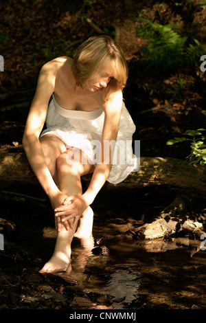 junge Frau sitzt auf einem Baumstamm an einem Bach durch einen Wald, kühlen ihre Füße, Deutschland Stockfoto