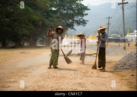 Eine Gruppe von Frauen, die eine staubige Bergstraße mit Strohbesen in der Nähe religiöser Strukturen und Steinkieshaufen in Taunggyi, Bundesstaat Shan, fegen. Myanmar Stockfoto