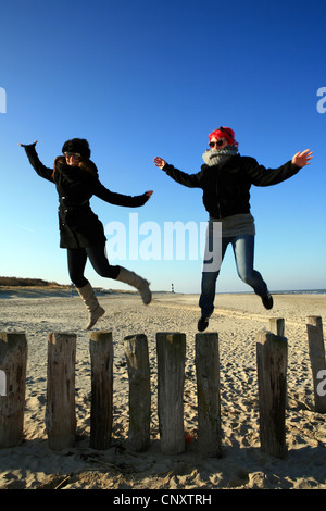 zwei junge Frauen frolicly springen von Buhnen am Strand, Niederlande, Zeeland, Breskens, Sluis Stockfoto