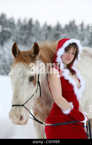 junge Frau in einem aufschlussreichen Santa Kostüm lehnte sich gegen ein weißes Pferd in einer Winterlandschaft Stockfoto