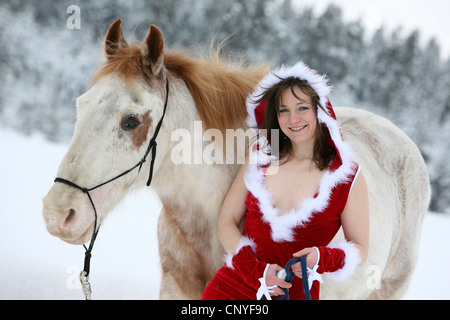 junge Frau in einem aufschlussreichen Santa Kostüm lehnte sich gegen ein weißes Pferd in einer Winterlandschaft Stockfoto