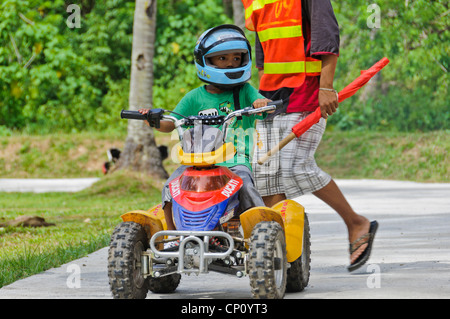 Kleine asiatische junge mit großen Helm Reiten Kinder ATV Quad beobachtet von Streckenposten mit roten Fahne - Puerto Galera, Philippinen Stockfoto