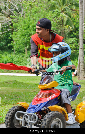 Kleine asiatische junge mit großen Helm Reiten Kinder ATV Quad unterstützt durch Streckenposten mit roten Fahne - Puerto Galera, Philippinen Stockfoto